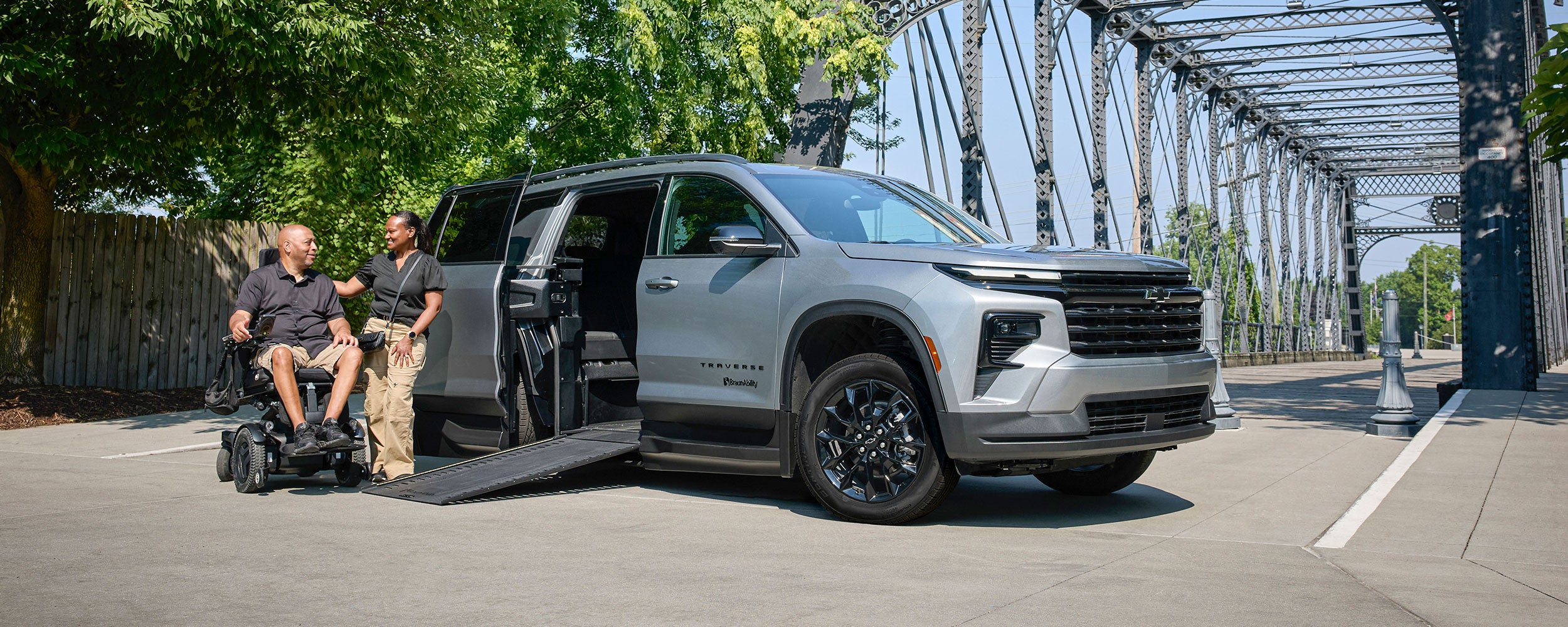 A Silver Chevy Traverse SUV With an Extended Wheelchair Ramp Parked on a Road Beside a Steel Bridge While Two People Converse Near the Ramp.